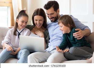 Happy Family Having Fun With Laptop At Home Together, Sitting On Cozy Couch, Smiling Young Mother And Father With Little Son And Daughter, Looking At Computer Screen, Shopping, Watching Movie Online