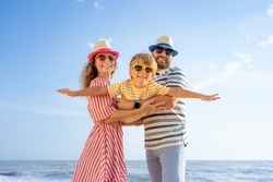 Happy Family Having Fun On The Beach. Mother And Father Holding Son Against Blue Sea And Sky Background. Summer Vacation Concept