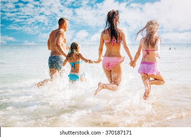 Happy Family Having Fun On The Beach. They With Holding Hands Running And Splashing In The Sea.