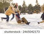 Happy family having fun sledding their smiling father together in winter park. Young parents with two children walking together outdoors in forest. Family leisure and winter holidays concept.