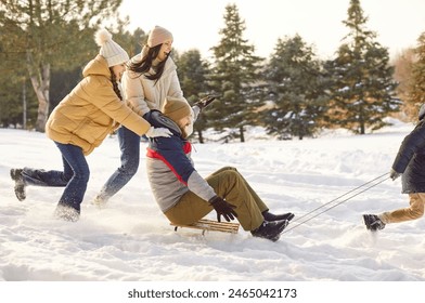 Happy family having fun sledding their smiling father together in winter park. Young parents with two children walking together outdoors in forest. Family leisure and winter holidays concept.