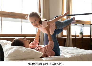 Happy Family Healthy Cheerful Young Mum Lifting Cute Little Child Daughter Up Playing Plane Doing Gymnastic Exercise In Bedroom, Funny Kid Girl Flying In Mothers Arms Look At Camera Having Fun On Bed