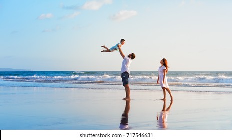 Happy family holidays. Joyful father, mother, baby son walk with fun along edge of sunset sea surf on black sand beach. Active parents and people outdoor activity on summer vacations with children.