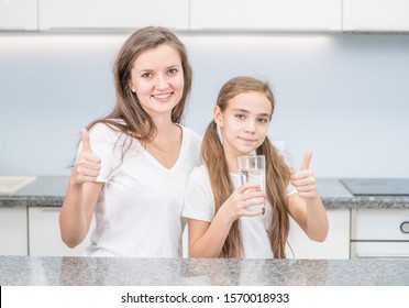 Happy Family At Home. Little Girl And Happy Mother With A Glass Of Water At Kitchen Show Thumbs Up Gesture