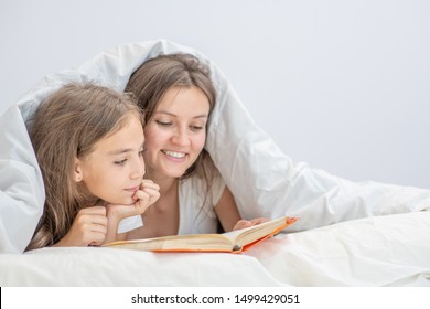 Happy family at home. Mother  and little girl reads a book on the bed under the blanket. Empty space for text