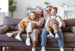 Happy Family Joyful Little Children Hugging Embracing With Positive Senior Grandparents While Sitting Together On Sofa In Living Room At Home, Cheerful Grandma And Grandpa With Kids Smiling At Camera