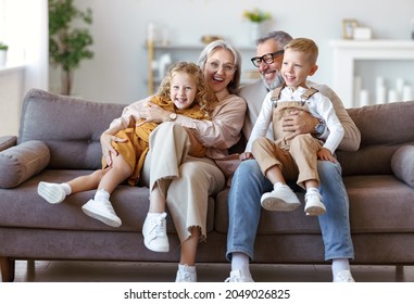 Happy family joyful little children hugging embracing with positive senior grandparents while sitting together on sofa in living room at home, cheerful grandma and grandpa with kids smiling at camera