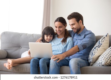 Happy Family With Kid Daughter Enjoy Using Laptop Apps, Making Online Call Together, Smiling Parents And Child Girl Relaxing Having Fun Watching Video On Computer, Doing Internet Shopping At Home