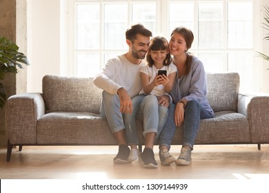 Happy Family And Kid Daughter Having Fun With Smartphone Gadget At Home, Little Child Girl Looking At Phone Play Game Using App With Mom Dad Watching Funny Mobile Video, Making Online Call On Couch