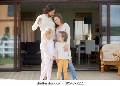 Happy Family With Kids Standing Outdoor Holding Keys Of Big Country House, Smiling Luxury Real Estate Owners Couple And Children Embracing On Terrace After Buying New Home, Mortgage Loan Concept