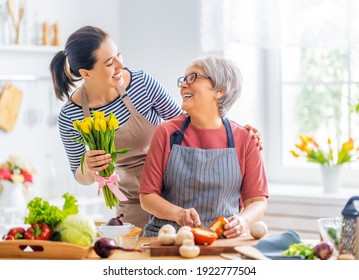 Happy Family In The Kitchen. Mother And Her Adult Daughter Are Preparing Proper Meal. Woman Is Giving Flowers To Her Mom.