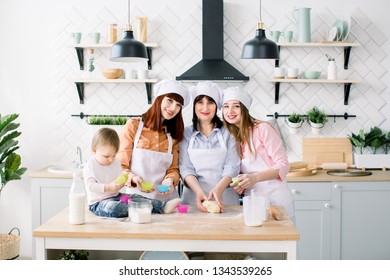 Happy Family In The Kitchen. Young Woman And Her Sister, Middle Aged Woman And Little Cute Daughter Cooking Cupcakes For Mothers Day, Casual Lifestyle Photo Series In Real Life Interior