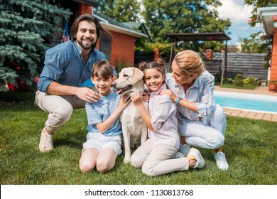 Happy Family With Labrador Dog Looking At Camera While Spending Time On Backyard Of Country House On Summer Day