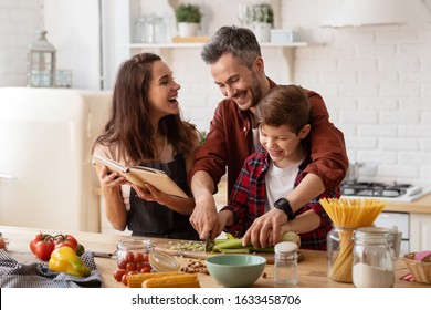 Happy Family Laughing Loud. Mother, Father And Preschool Son Cooking On Home Kitchen. Mom Holding Cookbook. Dad Helping Child To Chop Celery For Salad. Funny Weekends. Communication And Fun