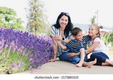 Happy Family Lifestyle And Holiday Concept. Mother, Little Boy, Girl Sitting, Walking In Old City, Street. Laughing On A Summer Sunny Day. Having Fun