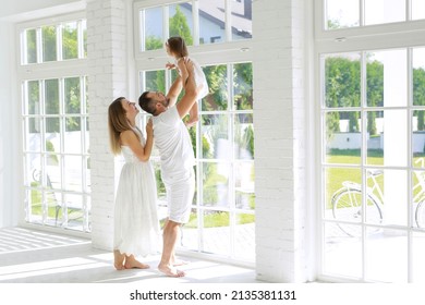 Happy family with a little daughter in a clean bright spacious apartment with large high windows. The father throws the child up. A sunny day for a family pastime together. Parents' white clothes - Shutterstock ID 2135381131