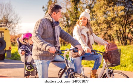 Happy Family With A Little Daughter Sitting On A Bike Seat Riding Bicycles By The Nature On A Sunny Winter Day