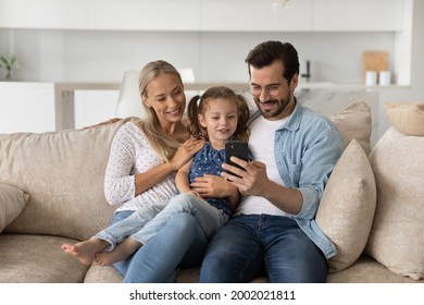 Happy Family With Little Daughter Using Smartphone Sitting On Couch, Smiling Mother And Father Hugging Adorable Preschool Child Girl, Looking At Phone Screen, Watching Video, Having Fun With Device