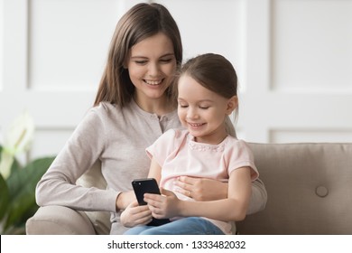 Happy Family Little Kid Daughter And Mom Holding Phone Having Fun With Smartphone, Young Mother With Child Girl Looking At Cellphone Enjoy Using Mobile Apps Play Games On Cellphone At Home On Sofa