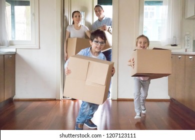 Happy Family With Little Kids Arrive At New Modern Home. Excited Funny Daughter And Son Holds Carton Boxes With Belongings Helps To Parents Running Inside House. Moving Relocation Day Mortgage Concept