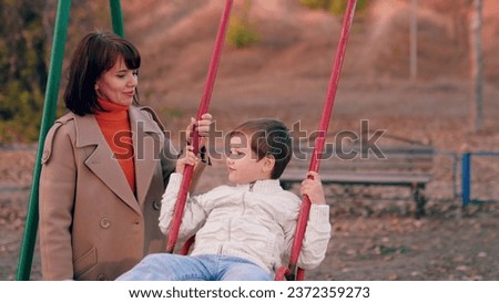 Happy family. Little son sitting on swing smiling, mom rocking child on children swing in park in fall. Autumn, little son plays on swing on playground under supervision of his mother. Mom kid play