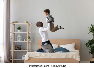 Happy Family Loving Black Father Holding Lifting Cute Small Mixed Race Child Son Playing On Bed, Funny Little Kid Boy Flying In Single African American Dad Arms Having Fun Feel Joy Bonding In Bedroom