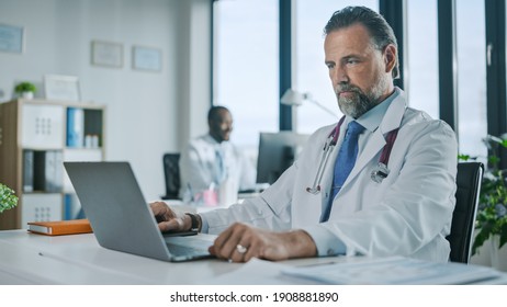 Happy Family Medical Doctor Is Working On A Laptop Computer In A Health Clinic. Physician In White Lab Coat Is Browsing Medical History Behind A Desk In Hospital Office.