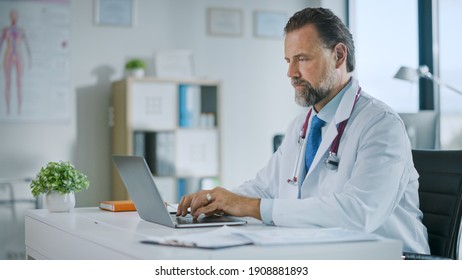 Happy Family Medical Doctor Is Working On A Laptop Computer In A Health Clinic. Physician In White Lab Coat Is Browsing Medical History Behind A Desk In Hospital Office.