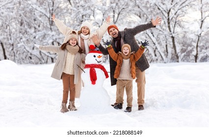 Happy family members dressed in warm clothes laughing merrily and raising hands up while posing around cheerful funny snowman in red hat and scarf, kids playing with parents in snowy park outdoors - Powered by Shutterstock