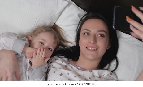 Happy Family Mom, Baby Take A Picture On Smartphone, Selfie Or Video Call To Dad Or Relatives In Bed. Photo Of Daughter's Mother And Child, Authenticity. New Generation, Always In Touch. Modern Gadget