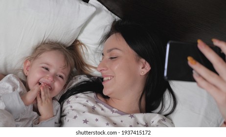 Happy Family Mom, Baby Take A Picture On Smartphone, Selfie Or Video Call To Dad Or Relatives In Bed. Photo Of Daughter's Mother And Child, Authenticity. New Generation, Always In Touch. Modern Gadget