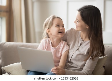 Happy Family Mom And Cute Kid Daughter Laughing Having Fun With Laptop Computer At Home Sitting On Sofa, Smiling Mother Looking At Little Child Girl Holding Pc Feeling Joy Enjoying Time Together