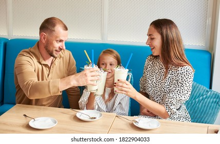 Happy Family - Mom, Dad And Daughter Are Sitting In The Local Cafe On A Blue Sofa Having Drinks, Clinking Glasses With Milkshakes. Plates And Cocktail Spoons On The Table