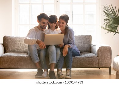 Happy family mom dad with kid daughter using laptop computer together on sofa, smiling parents and child girl watch funny video, do internet shopping, make online call on sunny day in living room