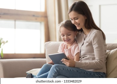 Happy family mom and kid daughter using digital tablet sitting on sofa, smiling parent mother with child girl holding pc computer looking at screen do online shopping make video call watch cartoons