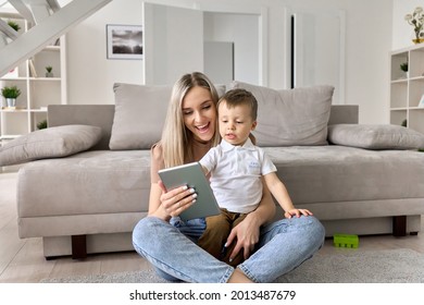 Happy family mom teaching cute child son holding digital tablet at home. Young mother and toddler kid boy using pad computer for playing, learning, watching funny videos or video call virtual meeting.