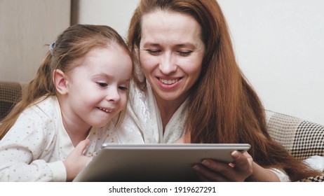 A happy family, mommy and a child are looking at an interesting cartoon on the tablet display and laughing, life is on online learning in the application, mom and daughter are lying on the couch and