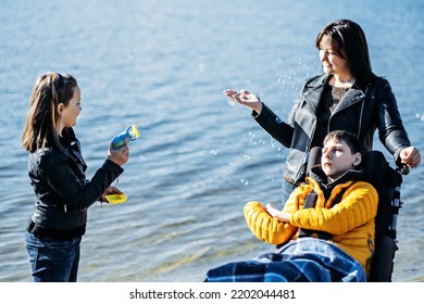 Happy Family, Mother, Daughter And Son With Cerebral Palsy Spending Time Together On The River Bank. Teen Boy Who Uses A Wheelchair Walking With Family