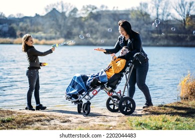 Happy Family, Mother, Daughter And Son With Cerebral Palsy Spending Time Together On The River Bank. Teen Boy Who Uses A Wheelchair Walking With Family