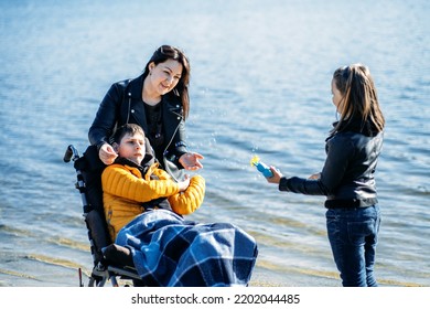 Happy Family, Mother, Daughter And Son With Cerebral Palsy Spending Time Together On The River Bank. Teen Boy Who Uses A Wheelchair Walking With Family