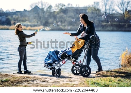 Happy family, mother, daughter and son with cerebral palsy spending time together on the river bank. Teen boy who uses a wheelchair walking with family
