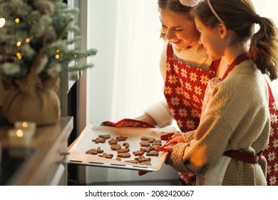 Happy Family Mother And Daughter Taking Freshly Baked Christmas Cookies From Oven While Cooking Together In Cozy Kitchen During Winter Holidays, Little Girl Helping Mom To Prepare For New Year At Home