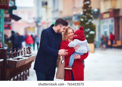 Happy Family, Mother, Father And Baby Walking Through The Winter Snowy City Street