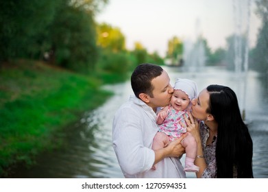 Happy Family Mother, Father, Child Daughter. Happy Young Family Spending Time Together Outside In Green Nature. Happy Family Having Fun Outdoor. Mom And Dad Kissing Their Baby