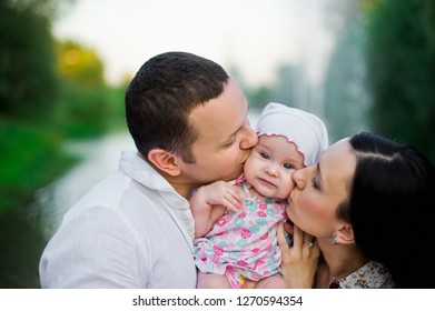 Happy Family Mother, Father, Child Daughter. Happy Young Family Spending Time Together Outside In Green Nature. Happy Family Having Fun Outdoor. Mom And Dad Kissing Their Baby