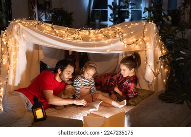 Happy family, mother, father and daughter lying inside self-made hut, tent in room in the evening and reading book. Winter coziness. Concept of fantasy, childhood, leisure time, love, care, home.
