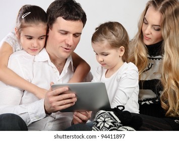 Happy Family Of Mother, Father And Daughters Sitting On A Sofa At Home Having Fun Using A Tablet Computer