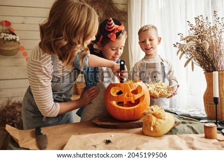 Happy family mother and kids carving pumpkin for Halloween holiday together, preparing for holiday party in kitchen, mom with little daughter and son smiling having fun while creating Jack-o-lantern