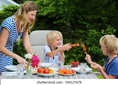 Happy Family, Mother With Kids, Two Funny Schoolboys, Laughing And Having Healthy Lunch Or Dinner Outside Sitting At Picnic Table In The Garden At The Backyard Of The House On A Sunny Summer Day