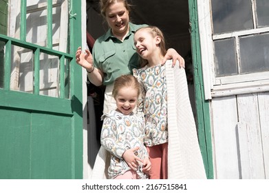 happy family mother with little kids daughters helper girls have fun in suburb countryside life on laundry day. candid mom hug and cover children with plaid and doing chores on porch of country house